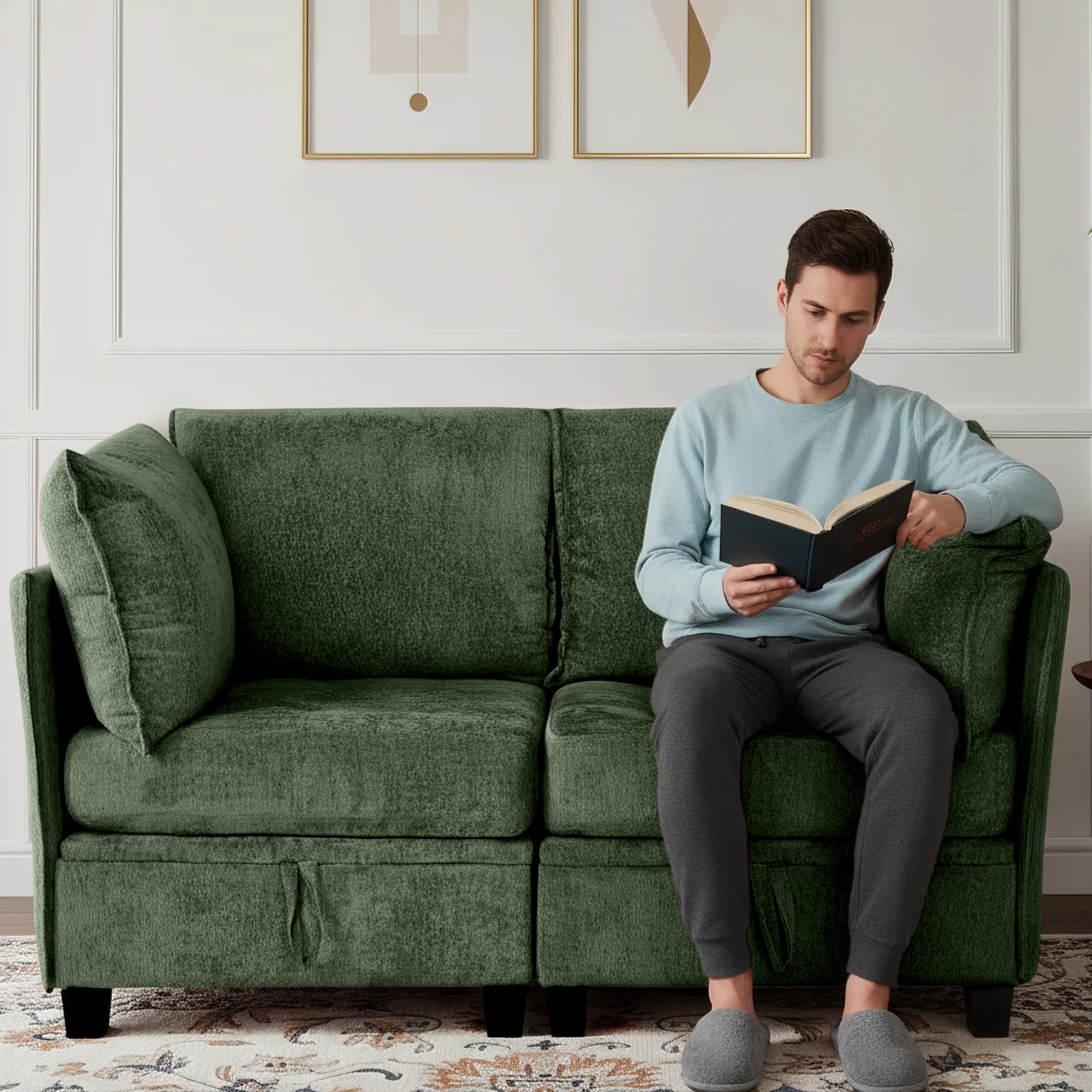 Man reading a book on a green sofa in a room with framed artwork on the wall.