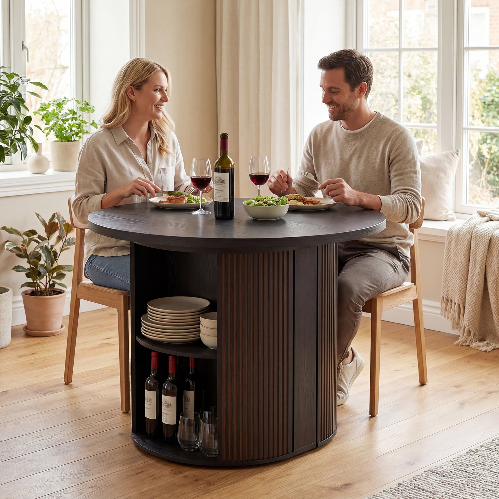Man and woman sitting at a round wooden table in a bright room with plants and wine bottles.